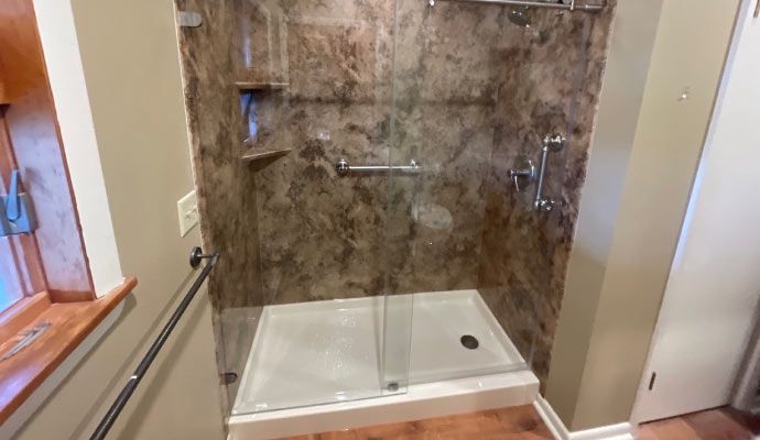 A completed walk-in shower featuring brown marble-patterned wall panels, a white low-profile base, glass doors, and a chrome safety grab bar.