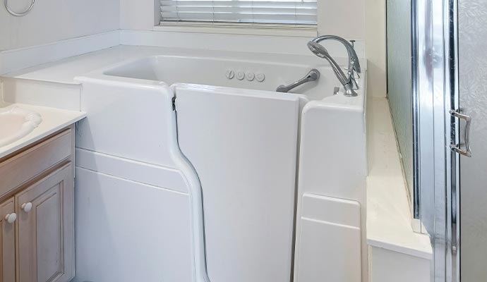 A white walk-in bathtub installed in a bathroom, showing the side entry door, a built-in seat, and a handheld shower faucet.